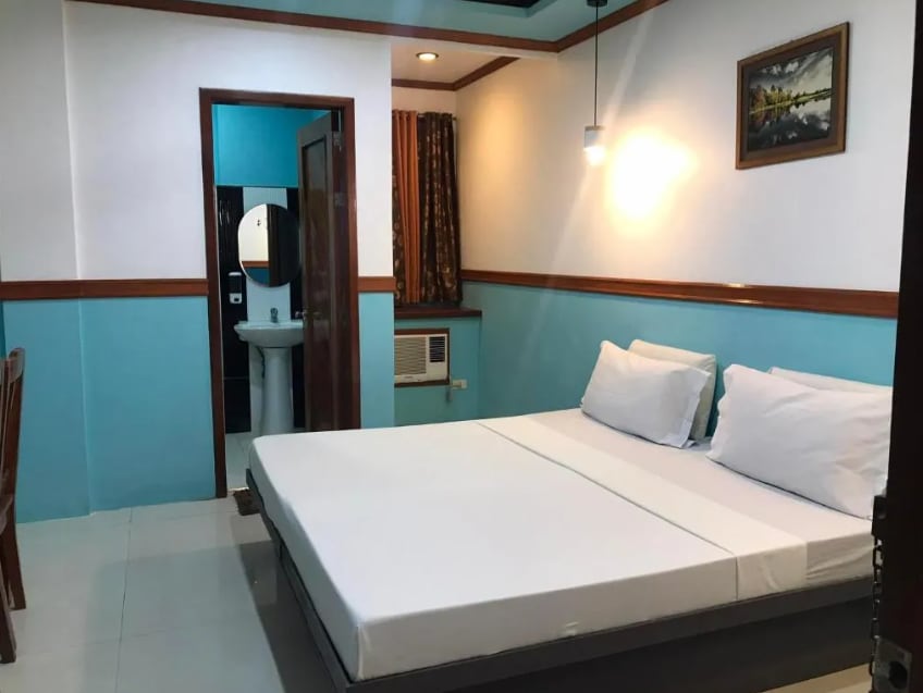 A standard room features a spacious bed with white linens and a simple headboard. A wooden chair and small table are positioned nearby. Bright turquoise walls are complemented by a framed artwork and warm lighting, creating a cozy ambiance. A private bathroom is accessible through a door.