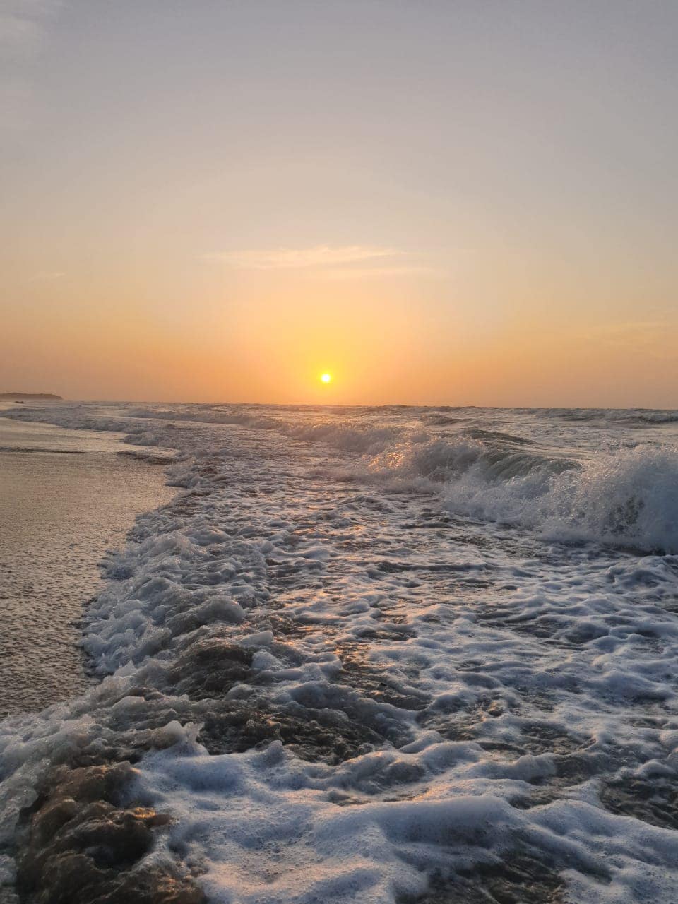 Waves gently break on a sandy shoreline as the sun sets on the horizon, casting warm orange and yellow hues across the sky. The water's surface is illuminated, with foam creating a dynamic texture along the edge of the beach.