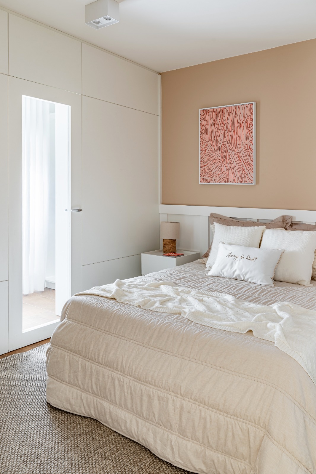 A well-lit bedroom features a spacious bed adorned with neutral-toned bedding and decorative pillows. A wall-mounted artwork adds a subtle pop of color. A closet with sliding doors and a nightstand with a lamp are visible, enhancing the room's functionality.