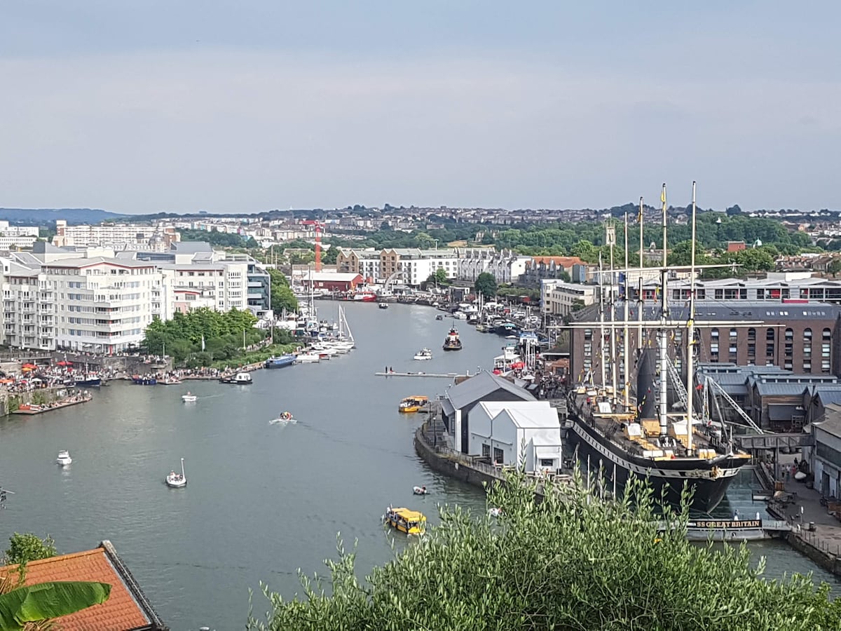 The image showcases a panoramic view of Bristol's floating harbour, with various boats traversing the water. A historic ship is docked alongside modern buildings, while lush greenery is visible in the foreground. The city skyline complements the scenic waterway.