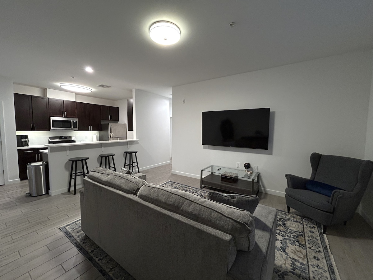 A modern living space features a comfortable gray sofa facing a large flat-screen television mounted on the wall. The kitchen is visible in the background, showcasing dark cabinetry and bar stools positioned at the counter. A stylish area rug adds softness to the room.