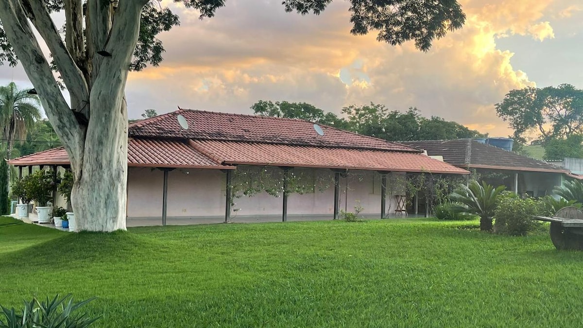 A spacious property is set amidst lush green grass, with a modest building featuring a red-tiled roof and multiple skylights. The landscape is accented by trees and potted plants, creating a tranquil outdoor environment during sunset.