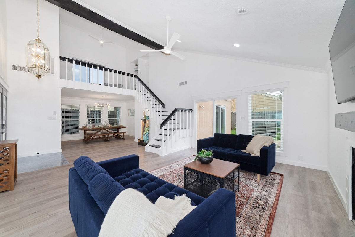 A spacious living area features two dark blue sofas facing a wooden coffee table. A light fixture hangs above, illuminating the room, while large windows provide natural light. A staircase leads to an upper level, offering a view of the dining area through glass doors.