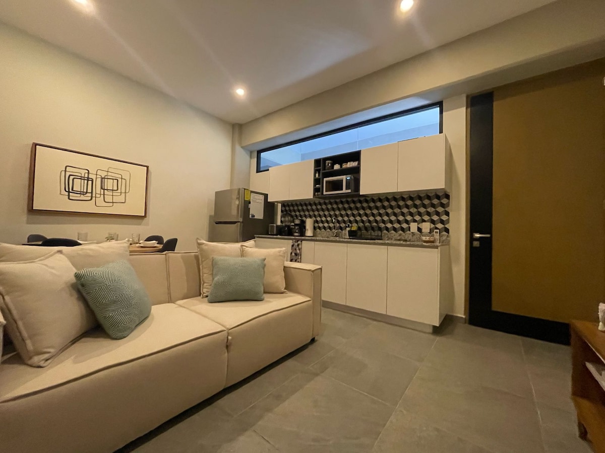 A modern kitchen area features sleek cabinetry and a patterned backsplash. An appliance station displays a microwave and coffee maker. A light-toned sofa with decorative cushions sits in the foreground, contributing to an open and inviting living space illuminated by soft overhead lighting.