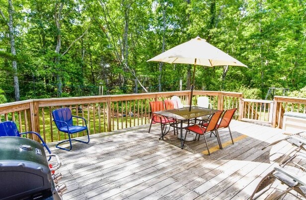 A spacious back deck is depicted, featuring a rectangular dining table surrounded by six colorful chairs. An umbrella provides shade over the table, and a grill is visible nearby. Lush greenery surrounds the area, enhancing the outdoor setting.