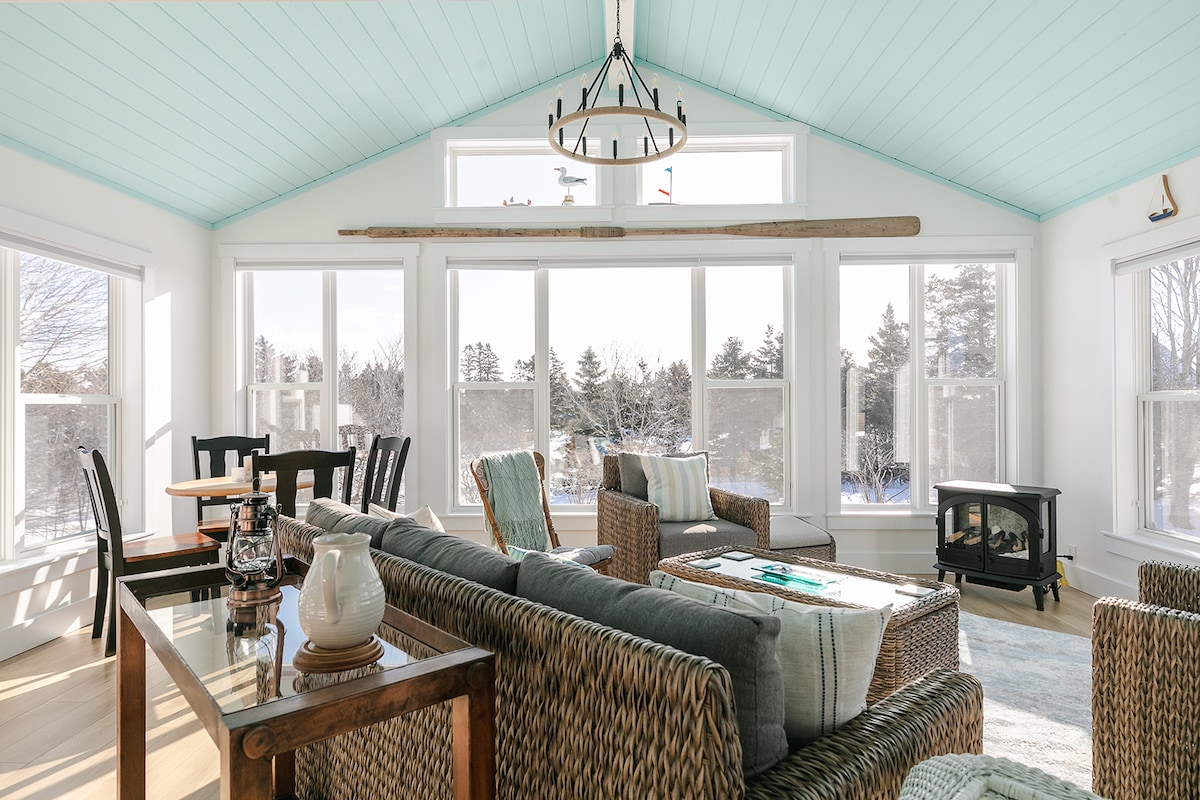 A sun-soaked sunroom features large windows with scenic views, allowing ample natural light to fill the space. Woven furniture is arranged for relaxation, complemented by a rustic chandelier. A wood-burning stove rests in the corner, enhancing the cozy ambiance of this inviting area.
