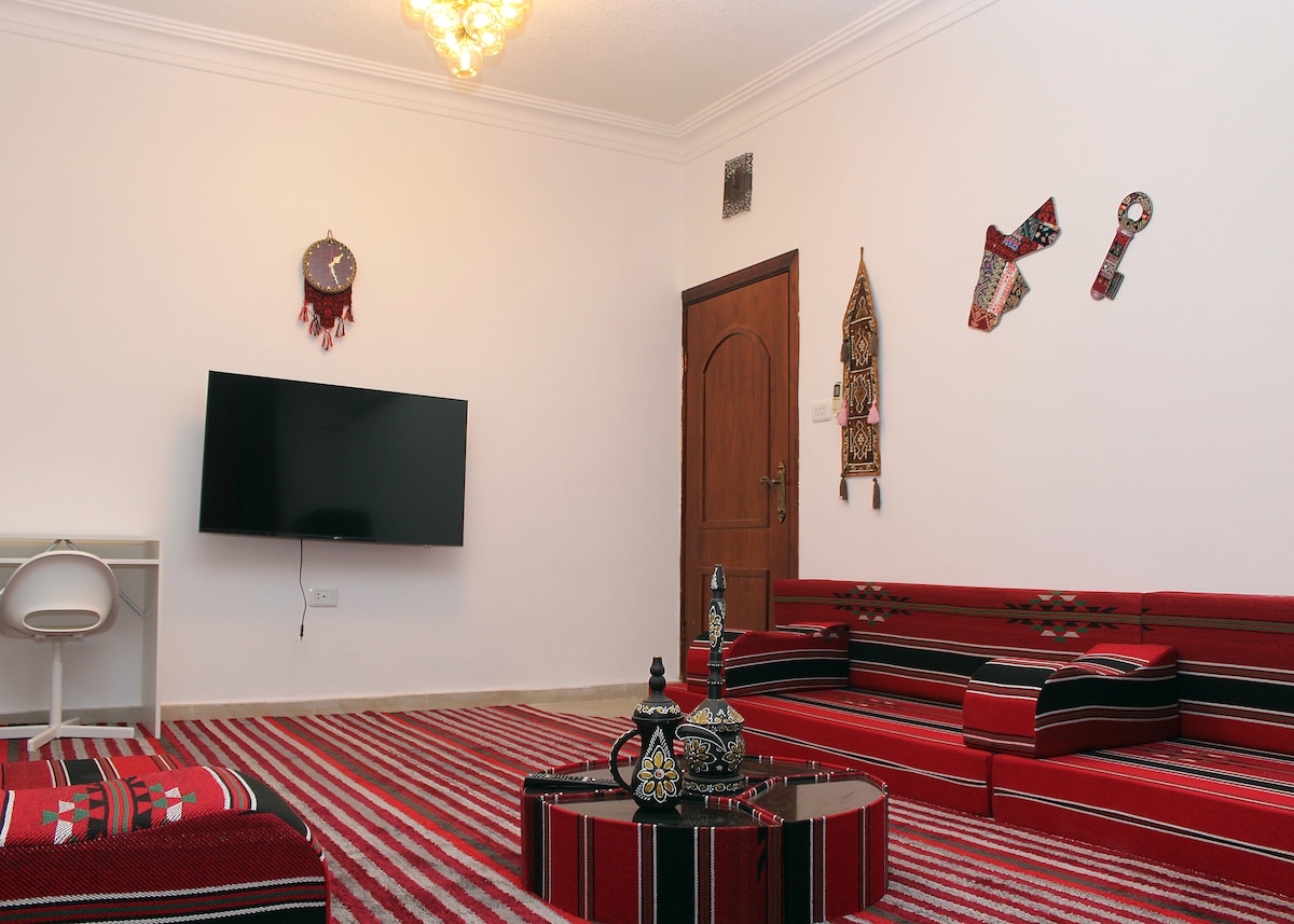 A living room features traditional Jordanian decor, including patterned seating along the walls and a low table. A flat-screen television is mounted on a wall opposite. Brightly colored wall hangings add cultural elements, while a stylish chandelier provides overhead lighting.