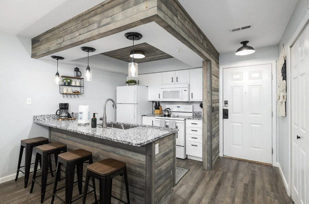 An updated kitchen area features a large granite countertop with seating for four, complemented by modern pendant lighting. A white refrigerator and stove are installed against a backdrop of wooden paneling, providing a contemporary yet warm space. The entry door is visible on the right.