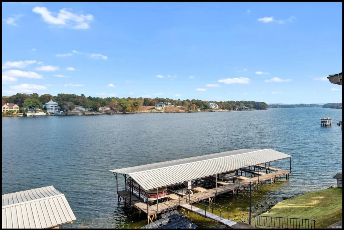 A scenic view of Lake Hamilton is captured from the balcony. The calm water reflects the blue sky and distant shoreline, lined with lush trees and residential homes. A boat slip is visible in the foreground, providing access to the water.