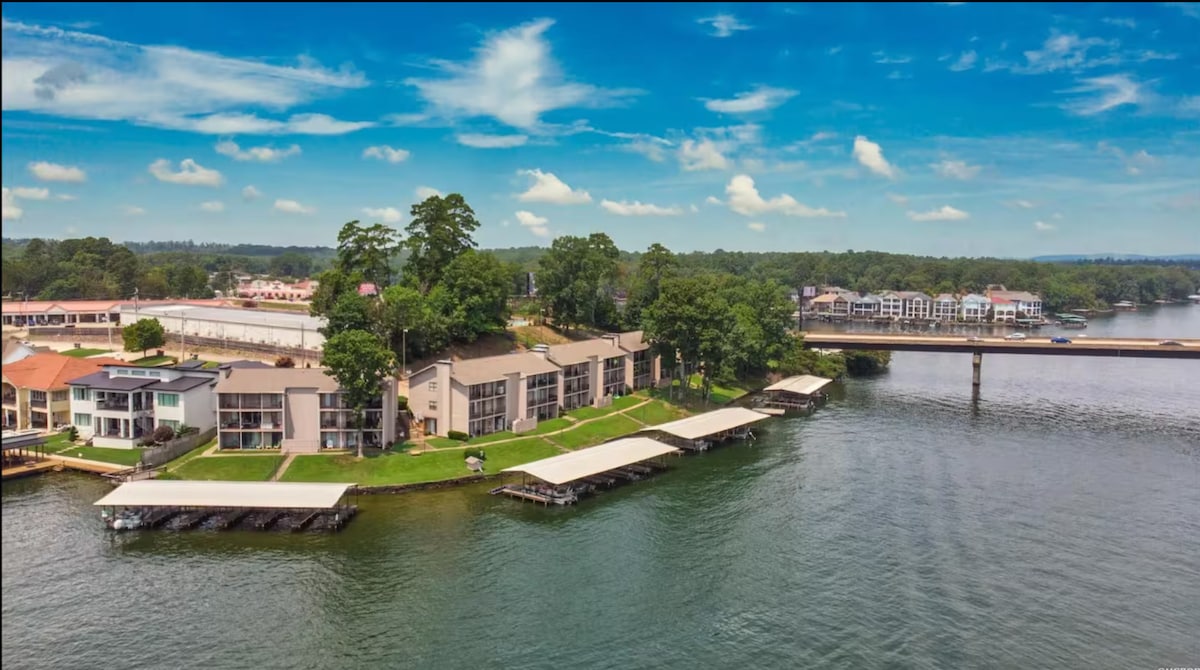 The aerial view captures the lakefront condo complex situated along Lake Hamilton. Boat slips are visible by the water's edge, with lush greenery surrounding the buildings. The scene is complemented by a blue sky dotted with light clouds.