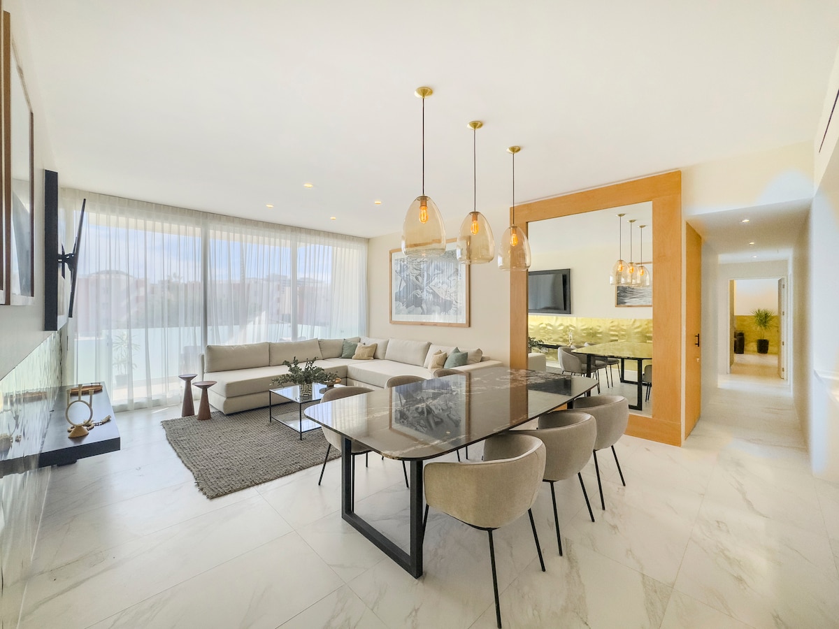 An open-plan living area features a modern design with large windows allowing natural light to fill the space. A dining table with soft, upholstered chairs is accompanied by a cozy seating arrangement. Elegant light fixtures hang above, and a textured rug adds warmth to the floor.