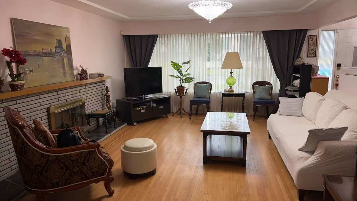 The living room is furnished with a white sofa and a patterned armchair, arranged around a central coffee table. A flat-screen television is positioned on a dark cabinet, with a decorative painting above a fireplace. Natural light filters through sheer curtains, complemented by a standing lamp and indoor plants.