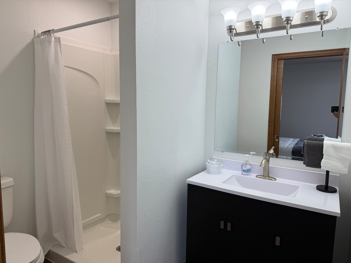 A clean and functional bathroom is showcased, featuring a walk-in shower with a shower curtain and built-in shelves. A sleek vanity with a white countertop and single basin sink is present, accompanied by modern lighting fixtures and a large mirror, enhancing the space's overall brightness.