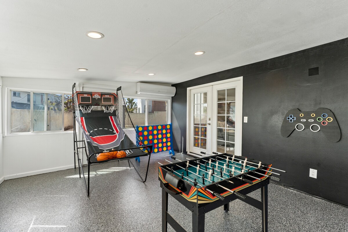 The game room features a range of entertainment options, including an indoor basketball arcade and a foosball table. Ample natural light shines through large windows, highlighting the vibrant wall art and the spacious layout. The flooring is covered with a durable surface, suitable for various activities.