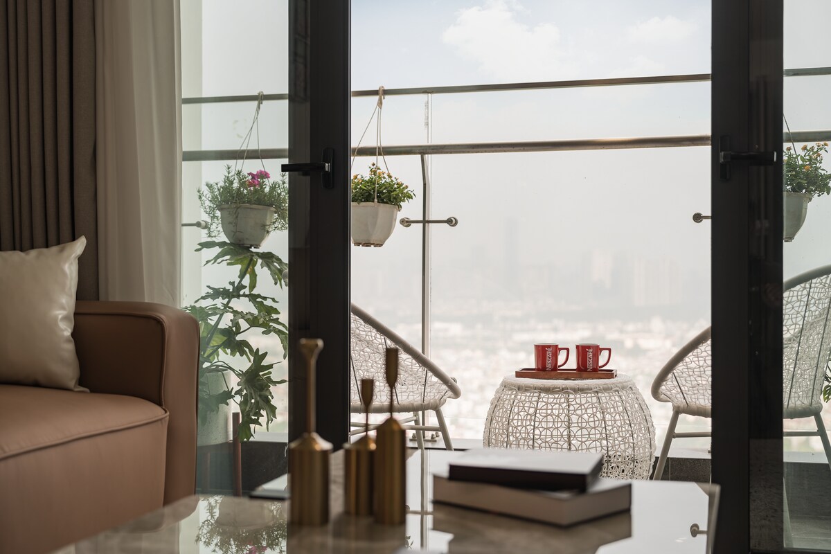 A cozy seating area is presented with a soft couch and a stylish coffee table, where two cups are positioned on a round side table. An outdoor balcony is visible through glass doors, showcasing hanging plants and two outdoor chairs, with a cityscape in the background.