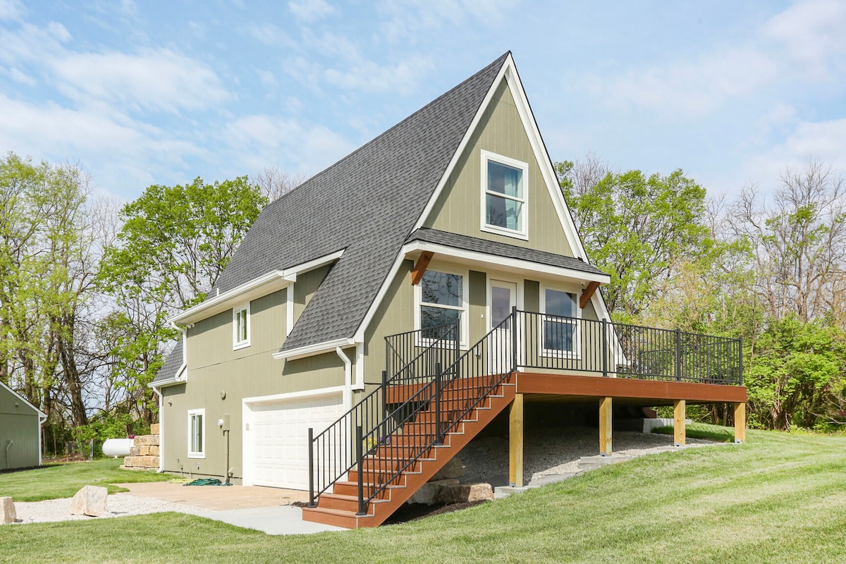 A modern A-frame home is showcased, featuring a striking green exterior and a sloped roof. A spacious deck with black railings is accessible by a set of stairs. The property is surrounded by landscaped grass and trees, contributing to a serene environment.