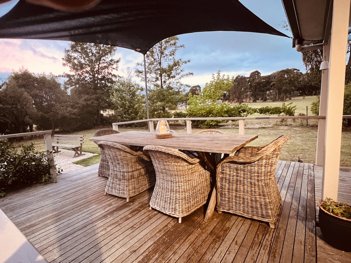 An outdoor dining area features a long wooden table surrounded by woven chairs, set under a shade canopy. Lush greenery and an open view of the garden enhance the relaxed atmosphere, with natural light filtering through the trees during twilight.