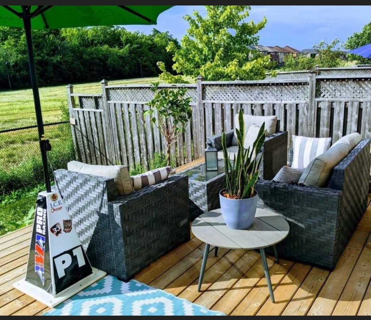 An outdoor seating area features two comfortable chairs, complemented by a low round table. A potted plant decorates the table, while a green umbrella provides shade. The space is surrounded by wooden fencing, offering a view of the greenery beyond.