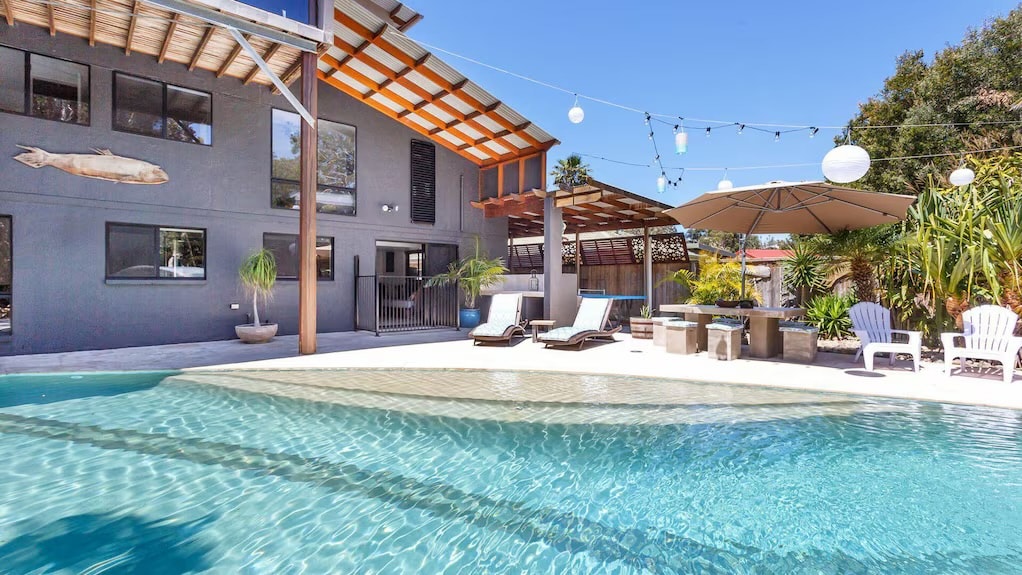 A serene outdoor area features a surfboard-shaped swimming pool with clear water reflecting the surroundings. Comfortable loungers are arranged beside the pool, and an umbrella provides shade for seating areas. Decorative lights hang above, enhancing the space's inviting feel.