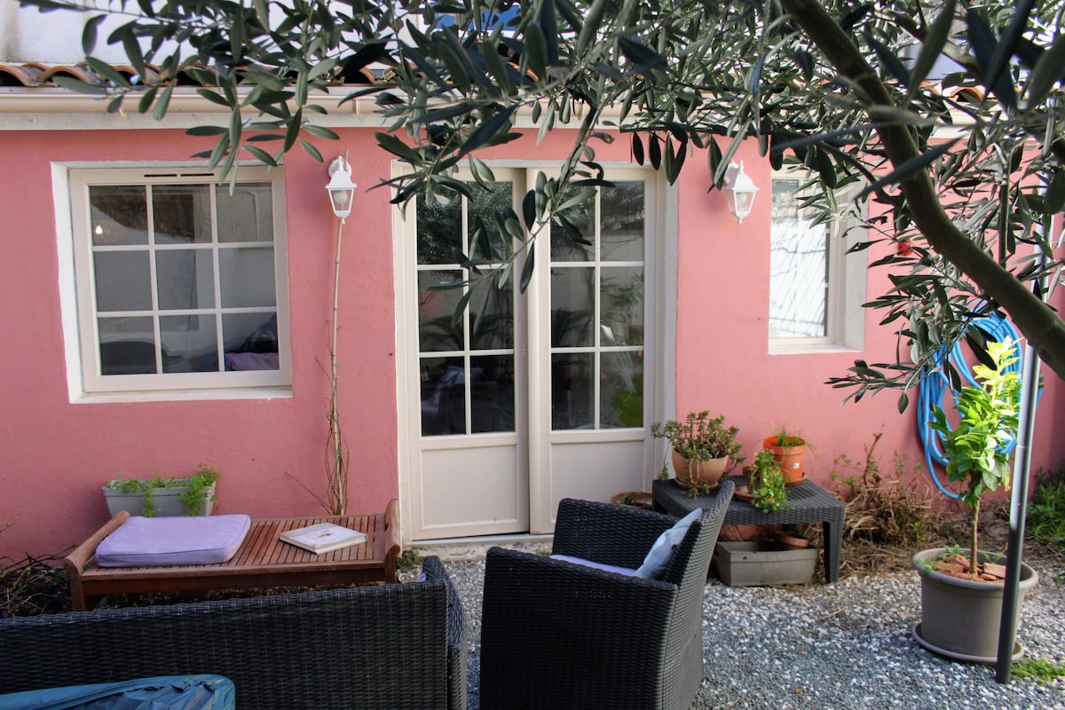 A charming exterior area is visible, featuring a pink facade with two glass-paneled doors. Outdoor seating includes wicker chairs and a small table amid potted plants. A tree provides shade, and there is a glimpse of greenery in the background.