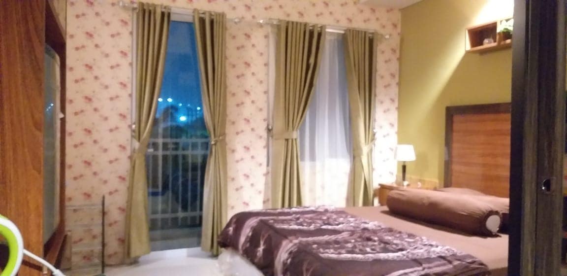A well-appointed bedroom features patterned wallpaper and two large windows with sheer and heavy drapes. A spacious bed is dressed in a cozy brown coverlet, complemented by bedside tables with lamps. Decorative shelves are mounted on the wall, adding a touch of character.