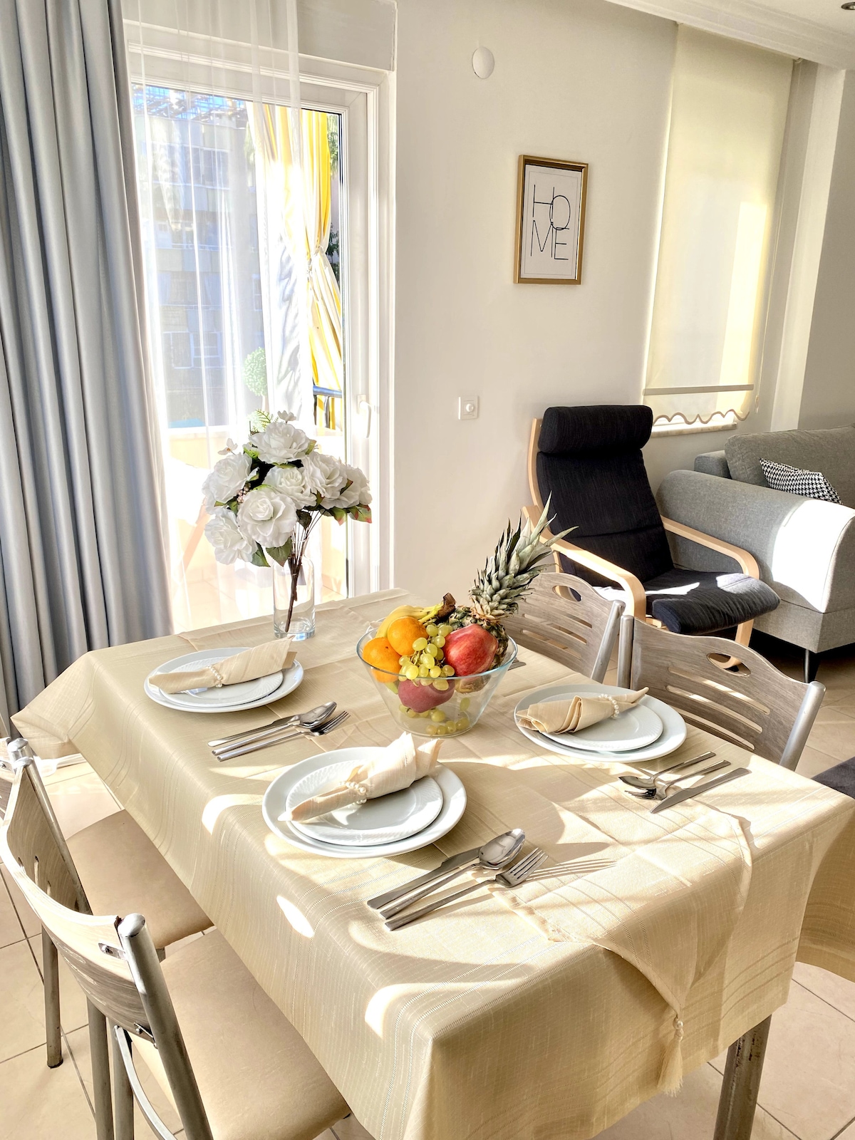 A dining table is set for four, featuring elegant plates and cutlery. A bowl filled with assorted fruit is centrally placed. Natural light streams in through a nearby window, enhancing the airy ambiance. A cozy seating area is visible in the background.