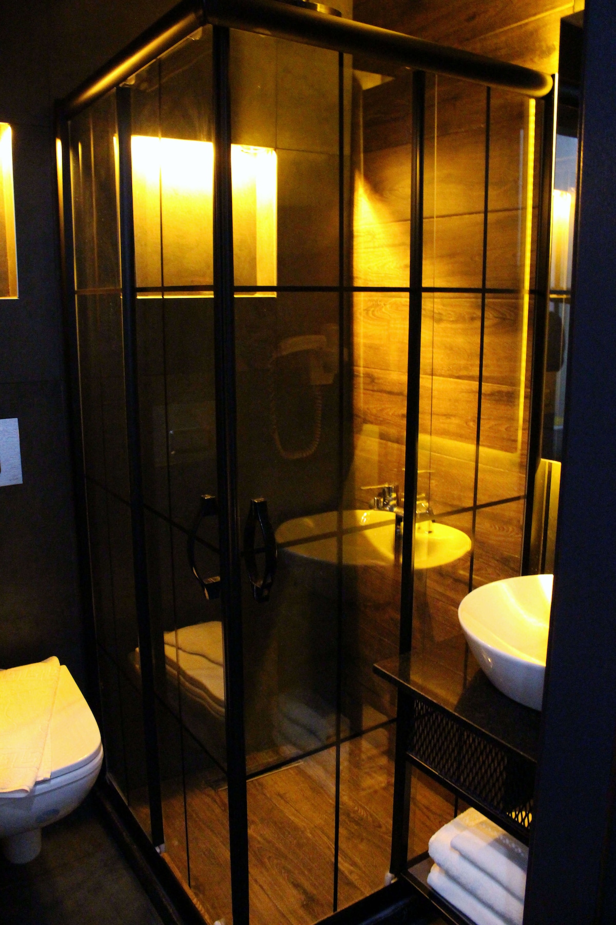 A modern bathroom is showcased, featuring a glass shower enclosure with black framing. Soft lighting casts a warm glow over the wooden accent walls. A compact sink with a round basin and a storage shelf with neatly arranged towels are also visible.