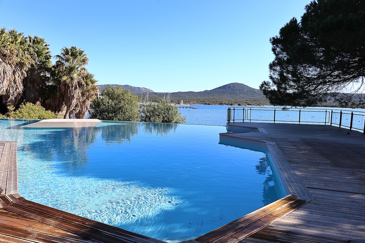 An infinity pool reflects the clear blue sky and calm waters nearby. Lush greenery and palm trees surround the area, providing a serene environment. The pool deck features wooden structures extending towards the water, contributing to a relaxing atmosphere.