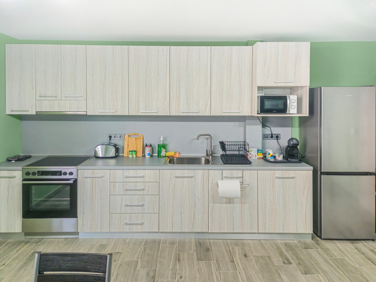 A modern kitchen is equipped with essential appliances, including an oven, stovetop, microwave, and refrigerator. Light wood cabinetry complements the green walls, while a stainless steel sink and countertops provide a functional workspace. Various kitchen items are neatly arranged on the counter.