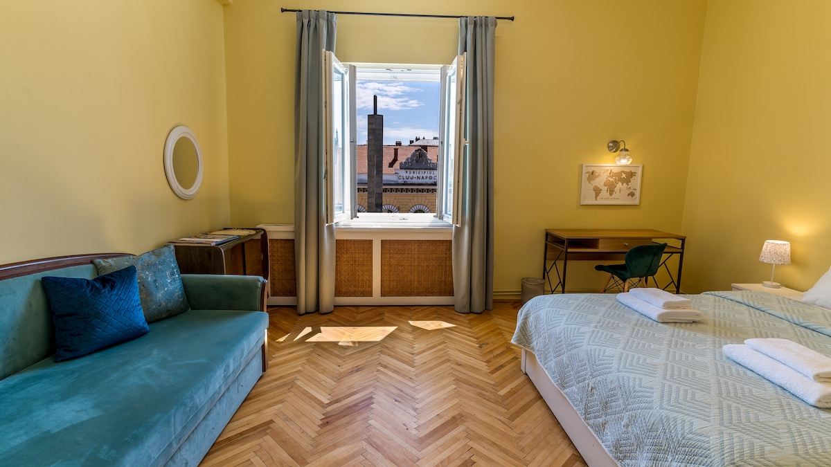 A spacious bedroom features a large queen bed with crisp linens and a cozy sofa positioned nearby. Natural light fills the room through a window adorned with light curtains, revealing glimpses of the cityscape outside. A dedicated work corner includes a simple desk and chair.
