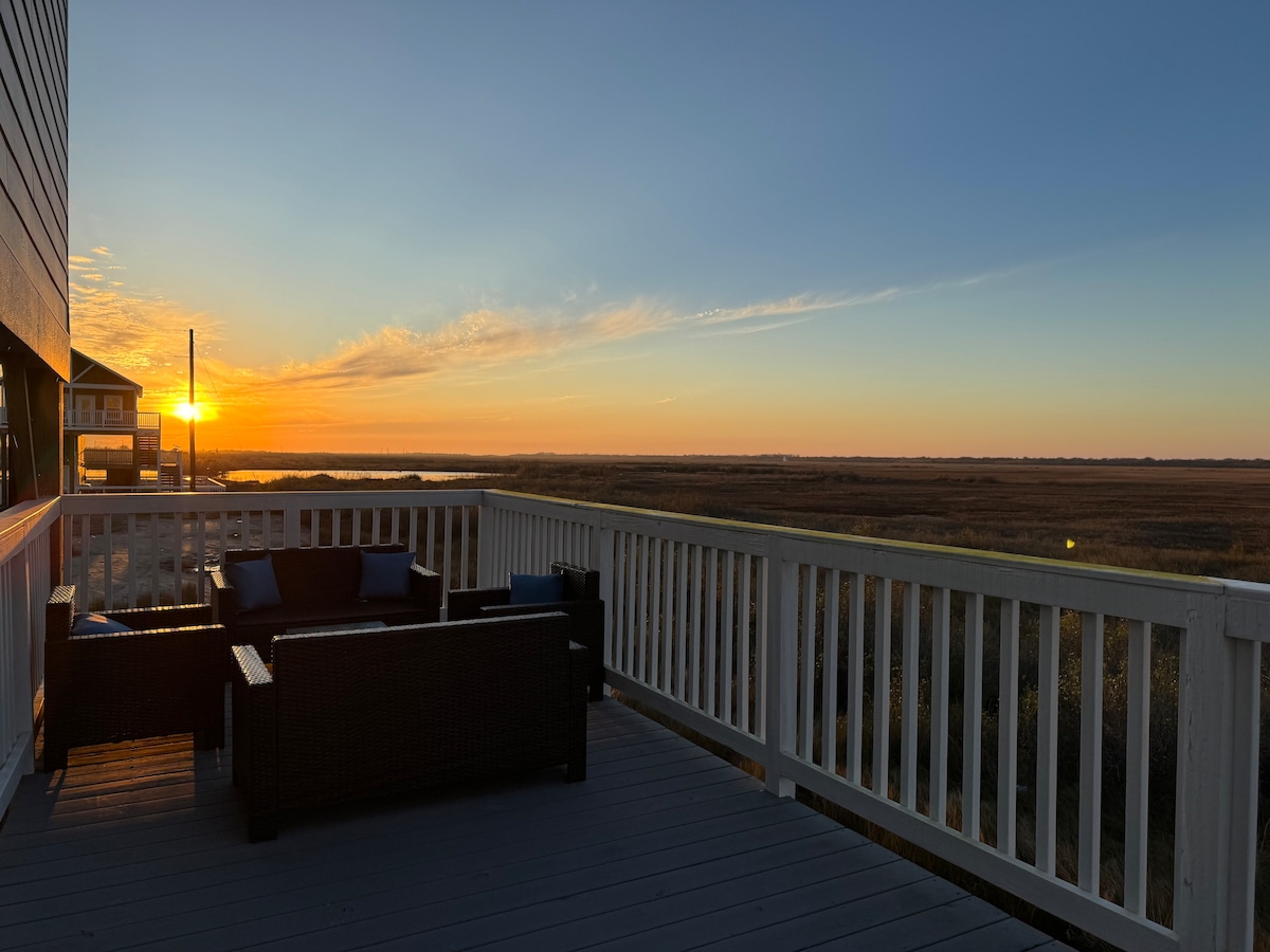 A spacious outdoor deck features comfortable seating arranged to take in the stunning sunset over the tranquil wetlands. The warm colors of the sky reflect on the calm water, creating a serene ambiance. The railing provides a clear view of the expansive landscape.