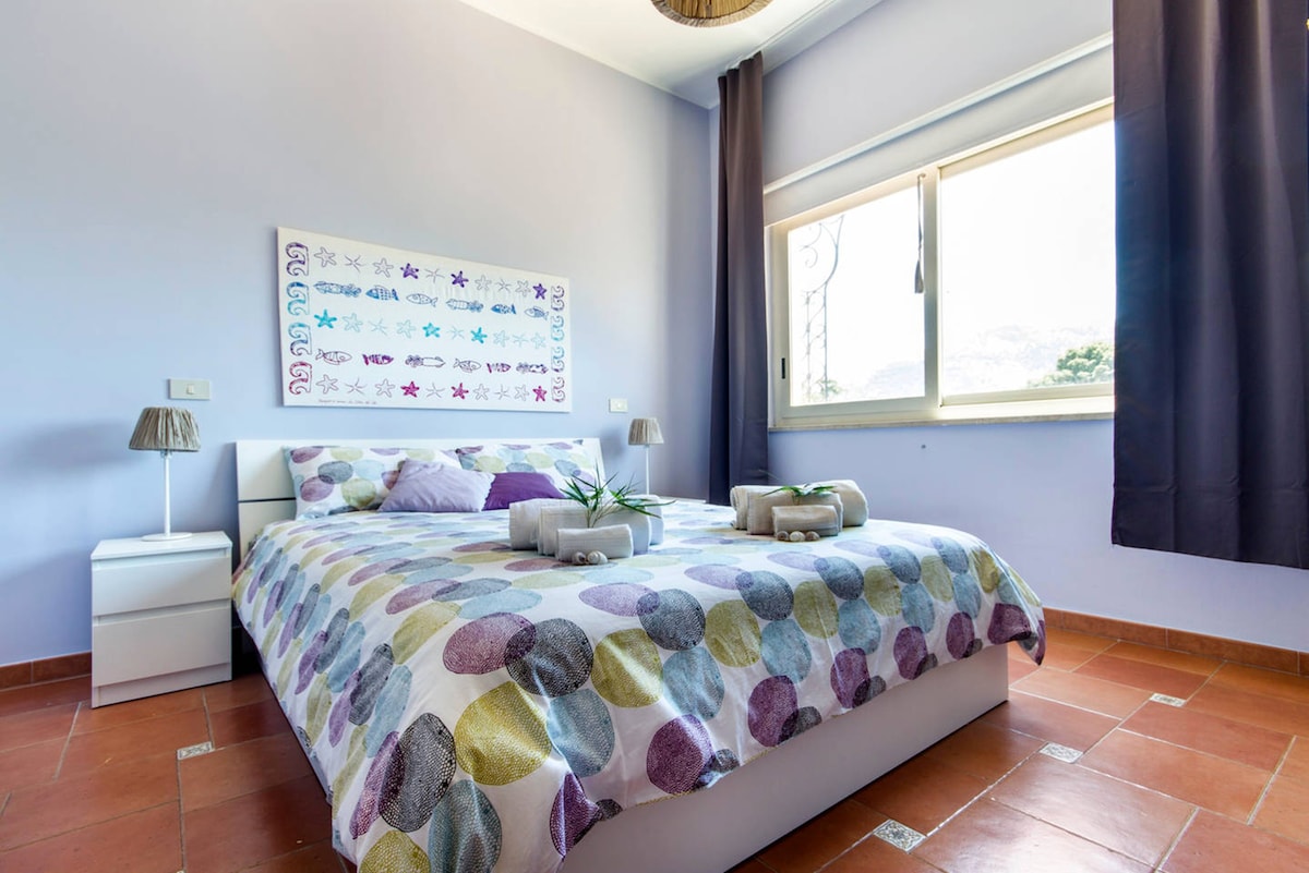 A well-lit bedroom features a spacious bed adorned with a multicolored patterned blanket. Two bedside lamps provide soft lighting, and a window allows natural light to fill the room, offering a view of the outdoors. Dark curtains frame the window.