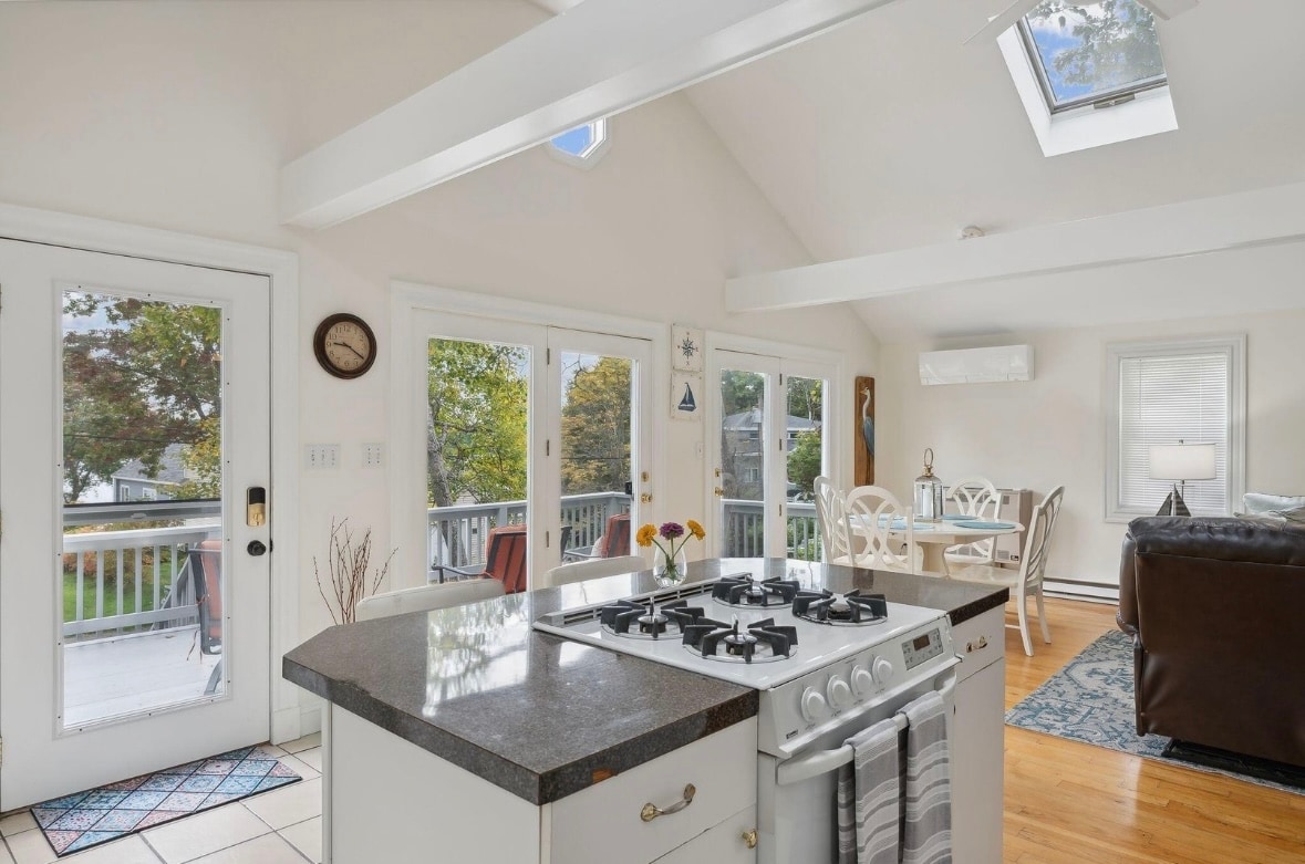 An open-concept kitchen features a gas stove with a black countertop, creating a functional space. Skylights above illuminate the area, while large glass doors provide access to the deck. A dining area and living room are visible, enhancing the bright and airy atmosphere.