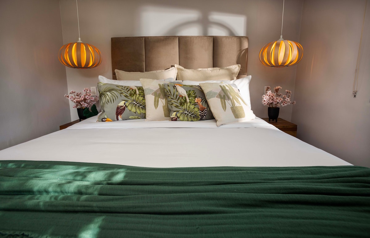 A cozy sleeping area features a spacious bed adorned with decorative pillows and a soft green throw. Two stylish pendant lights hang above, illuminating the design. Small potted plants are placed on either side of the bed, enhancing the tranquil atmosphere.