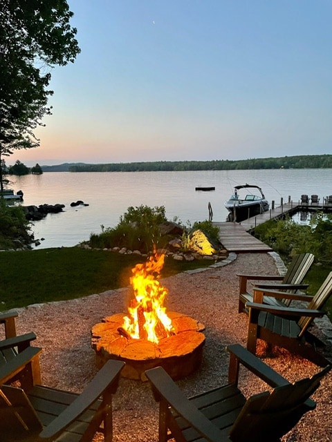 A fire pit surrounded by several wooden Adirondack chairs is set near the water's edge. The flickering flames provide warmth against the backdrop of a serene lake, while a boat is docked nearby. Lush greenery and rocks frame the picturesque scene as dusk approaches.