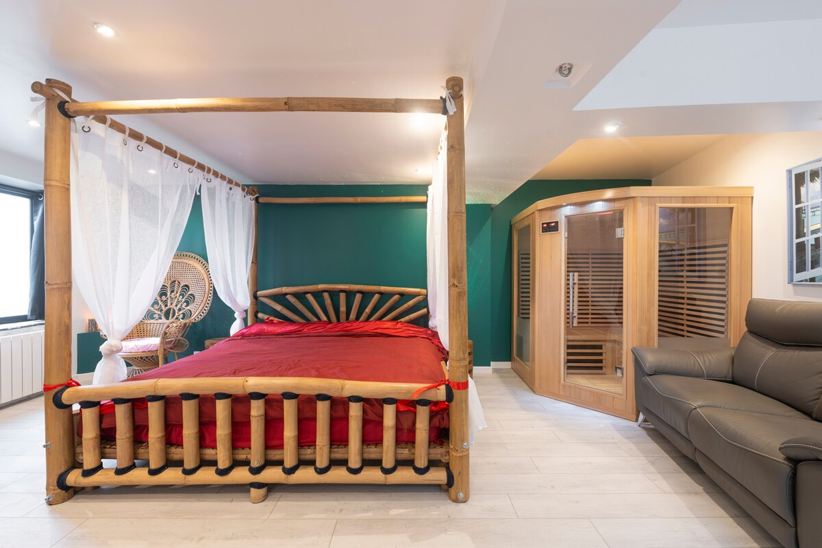 A spacious room features a wooden bed with a vibrant red coverlet, complemented by sheer white curtains. A modern sauna is visible nearby, alongside a comfortable seating area, where a full-length mirror adds depth to the space. A rattan chair provides an additional element of relaxation.