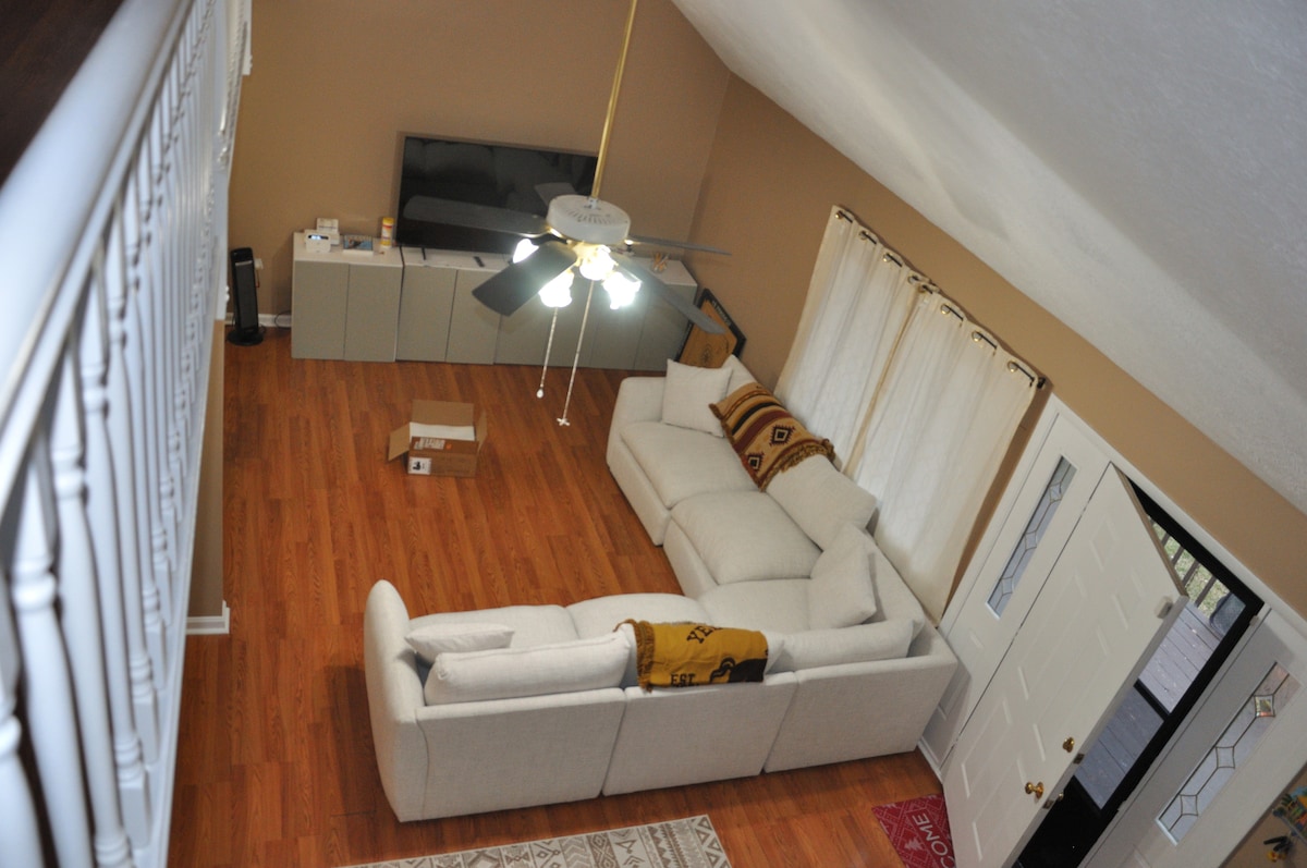 A spacious living area is presented, featuring a light-colored sectional sofa positioned near a wall-mounted television. Natural light enters through sheer curtains, and a ceiling fan provides comfort. A decorative area rug complements the hardwood flooring, while an entry door is visible in the background.