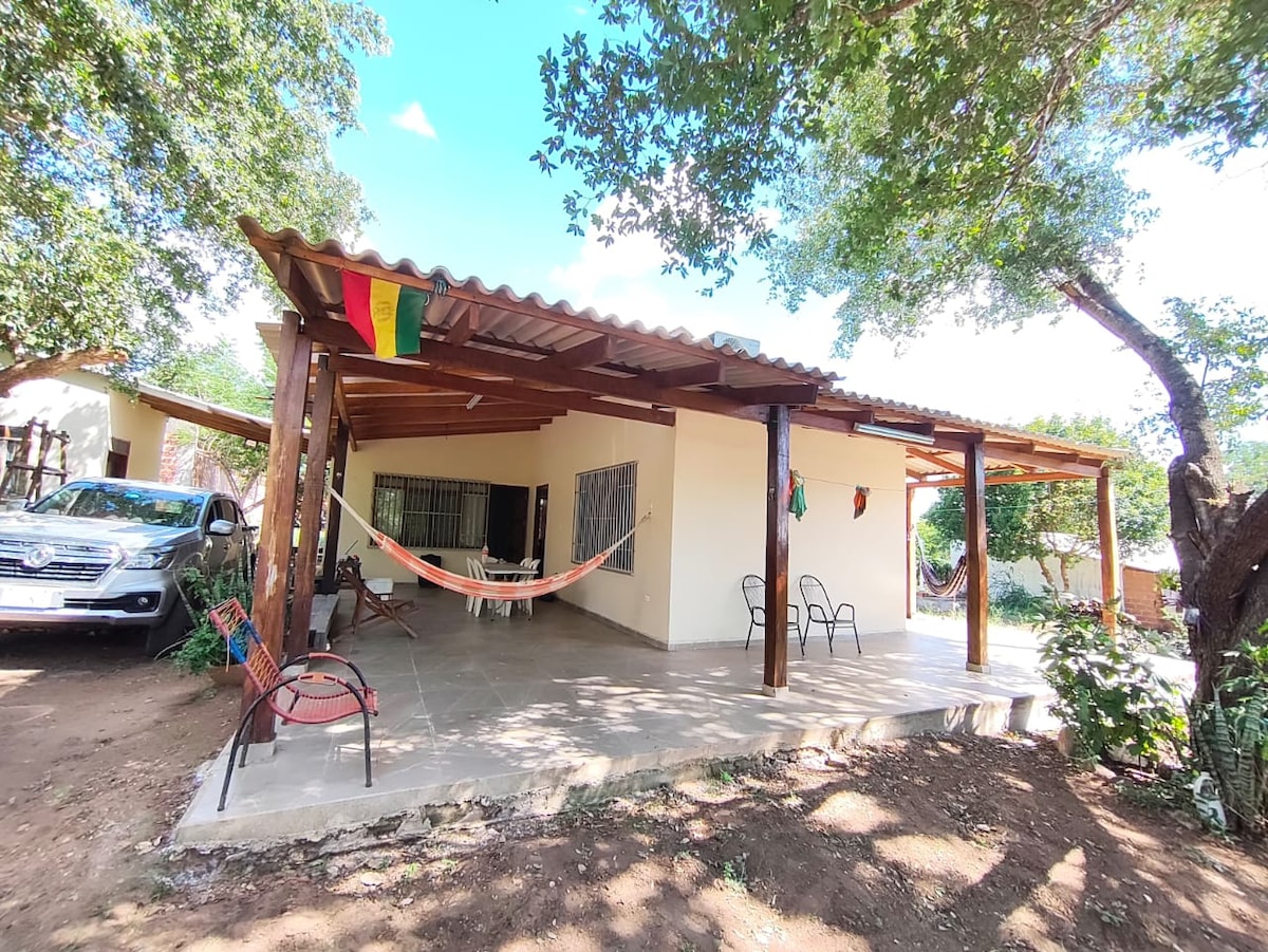 A covered outdoor area features wooden beams and a tiled floor, offering space for relaxation. Two hammocks are seen hanging, alongside a few chairs. Nearby, a vehicle is parked under the shade of trees, providing a comfortable and inviting setting.