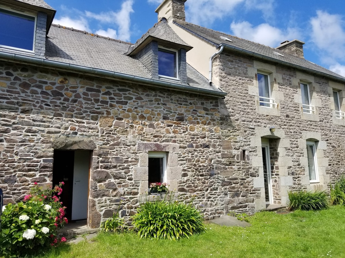A charming stone house is presented, showcasing a well-maintained facade adorned with greenery and seasonal flowers. Multiple windows allow ample natural light, while the expansive front lawn enhances the inviting atmosphere. The entrance is highlighted by a simple doorway flanked by colorful flower beds.