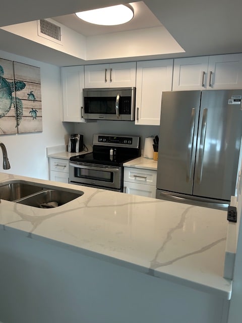 A modern kitchen features white cabinetry and sleek stainless steel appliances, including a microwave and refrigerator. The countertops display a marble-like pattern. A coffee maker is placed on the counter, alongside a painting of a turtle on the wall, adding a coastal touch.