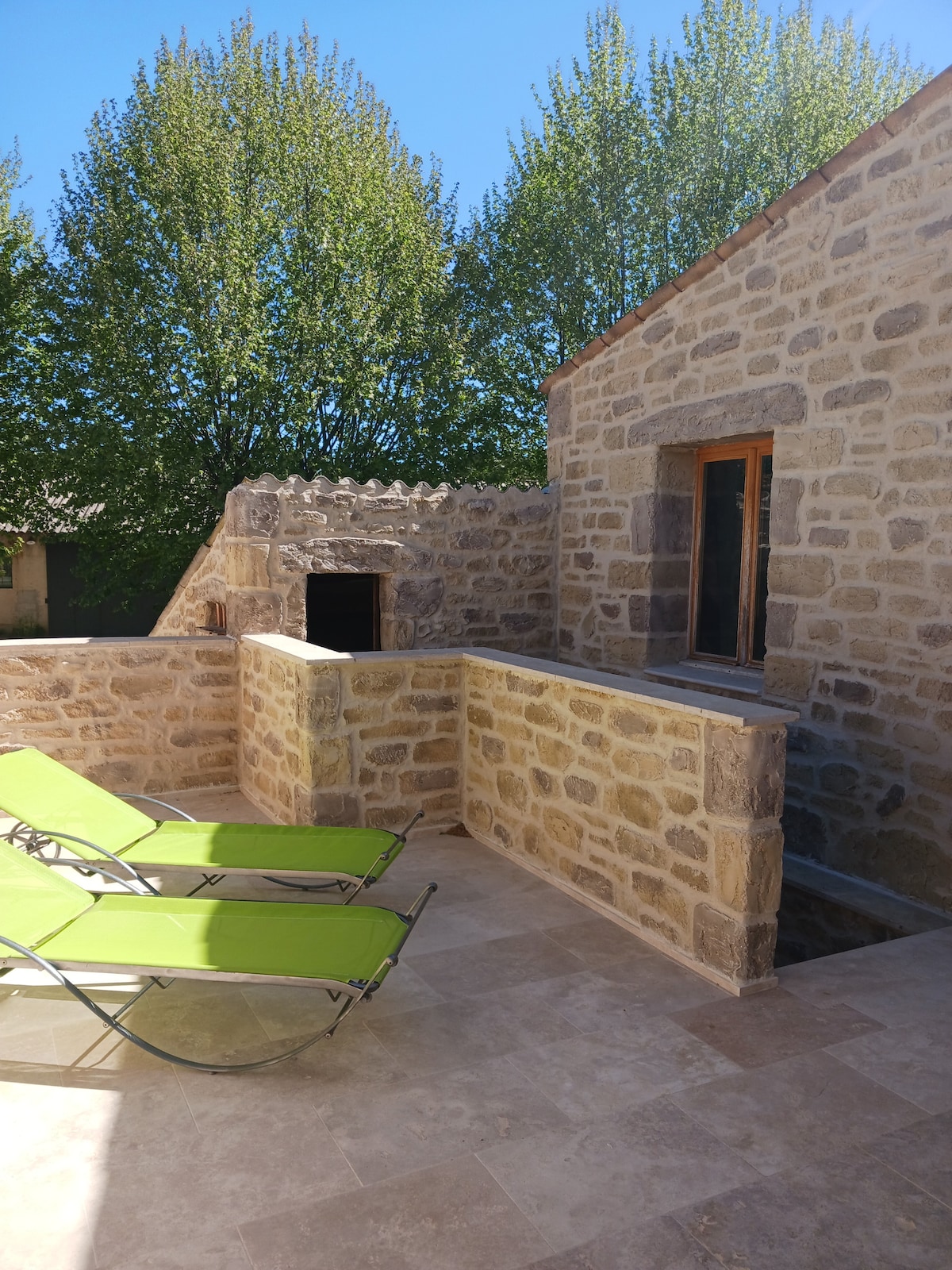 A sunlit terrace is enclosed by natural stone walls, featuring two green lounge chairs positioned for relaxation. Leafy trees stand in the background, contributing to a serene outdoor atmosphere. The texture of the stone complements the warm tones of the building, enhancing the setting.