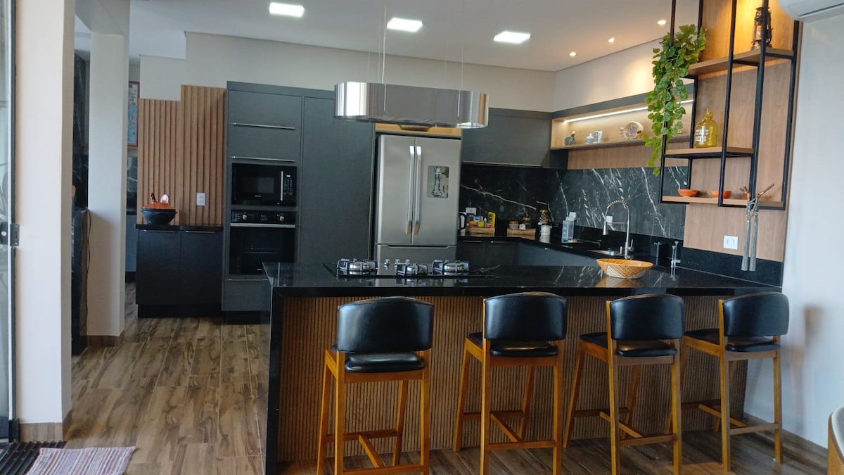 The modern kitchen features sleek black cabinetry and a large central island with four high-backed chairs. A gas stove, built-in oven, and stainless-steel refrigerator are visible, complemented by stylish marble countertops and open shelving displaying dishware.