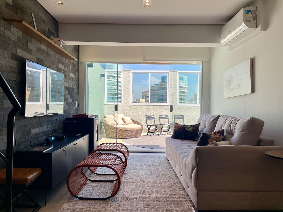 A cozy living area features a comfortable sofa facing a modern entertainment unit with a Smart TV. Large glass doors lead to a balcony, allowing natural light to fill the space. A rug adds texture, and the room is complemented by city views visible through the windows.