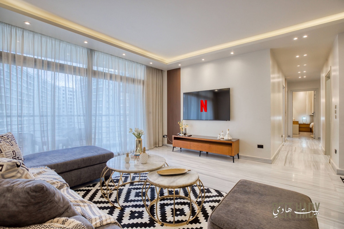 A modern living room features a large gray sectional sofa positioned around a circular coffee table on a patterned area rug. A wall-mounted TV showcases a streaming service interface, while natural light floods in through sheer curtains covering a balcony door.