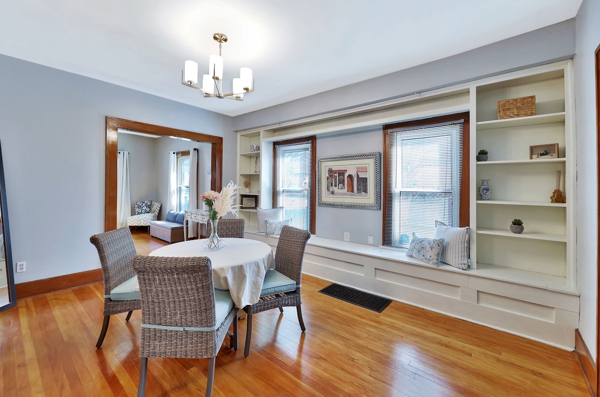 A dining area is furnished with a round table surrounded by four woven chairs. Large windows allow natural light to illuminate the space, and built-in shelves are visible in the background, showcasing a few decorative items. A mirror reflects part of the adjacent living room.
