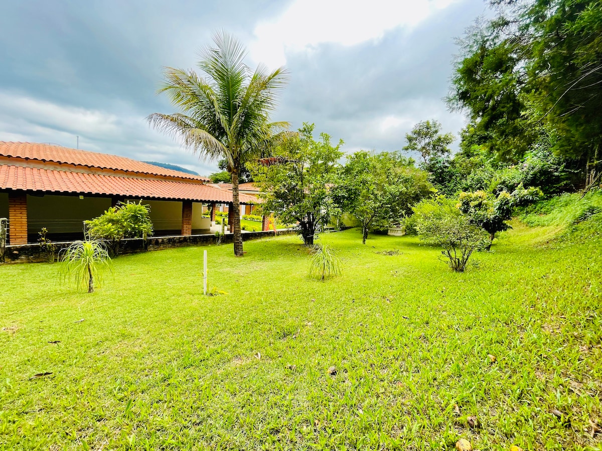 A spacious garden surrounds the house, featuring well-maintained grassy areas and diverse vegetation. A variety of trees and shrubs create a natural setting, while a palm tree stands prominently. The tranquil atmosphere highlights the connection to nature, inviting relaxation and exploration.