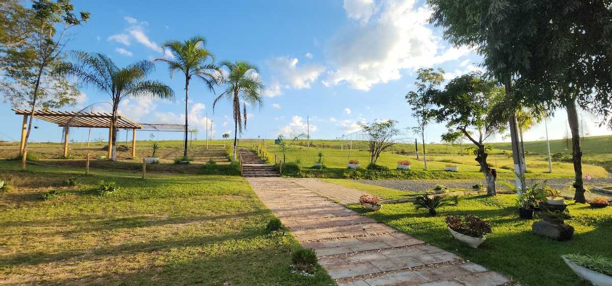 A sunlit pathway leads through a landscaped area featuring palm trees and greenery. A wooden gazebo is positioned to one side, while flower beds add color along the edges. Open fields stretch into the distance under a partly cloudy sky.