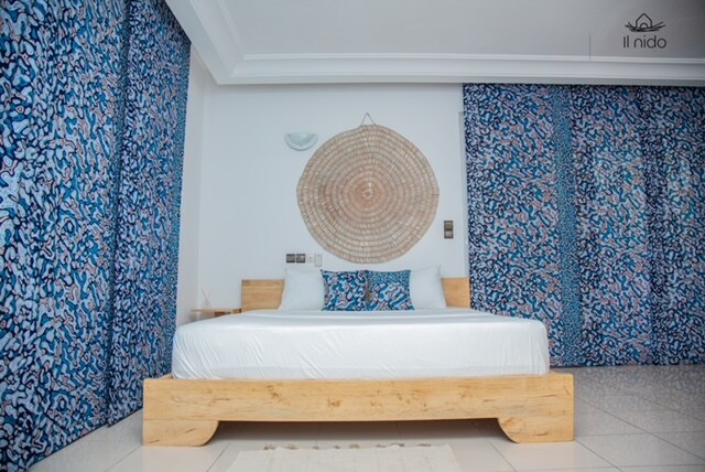 A spacious sleeping area features a light wooden bed with white bedding. Vibrant blue patterned walls provide a striking backdrop. Decorative circular wall art is positioned above the headboard, while additional patterned curtains frame the windows, allowing natural light to enhance the room's design.