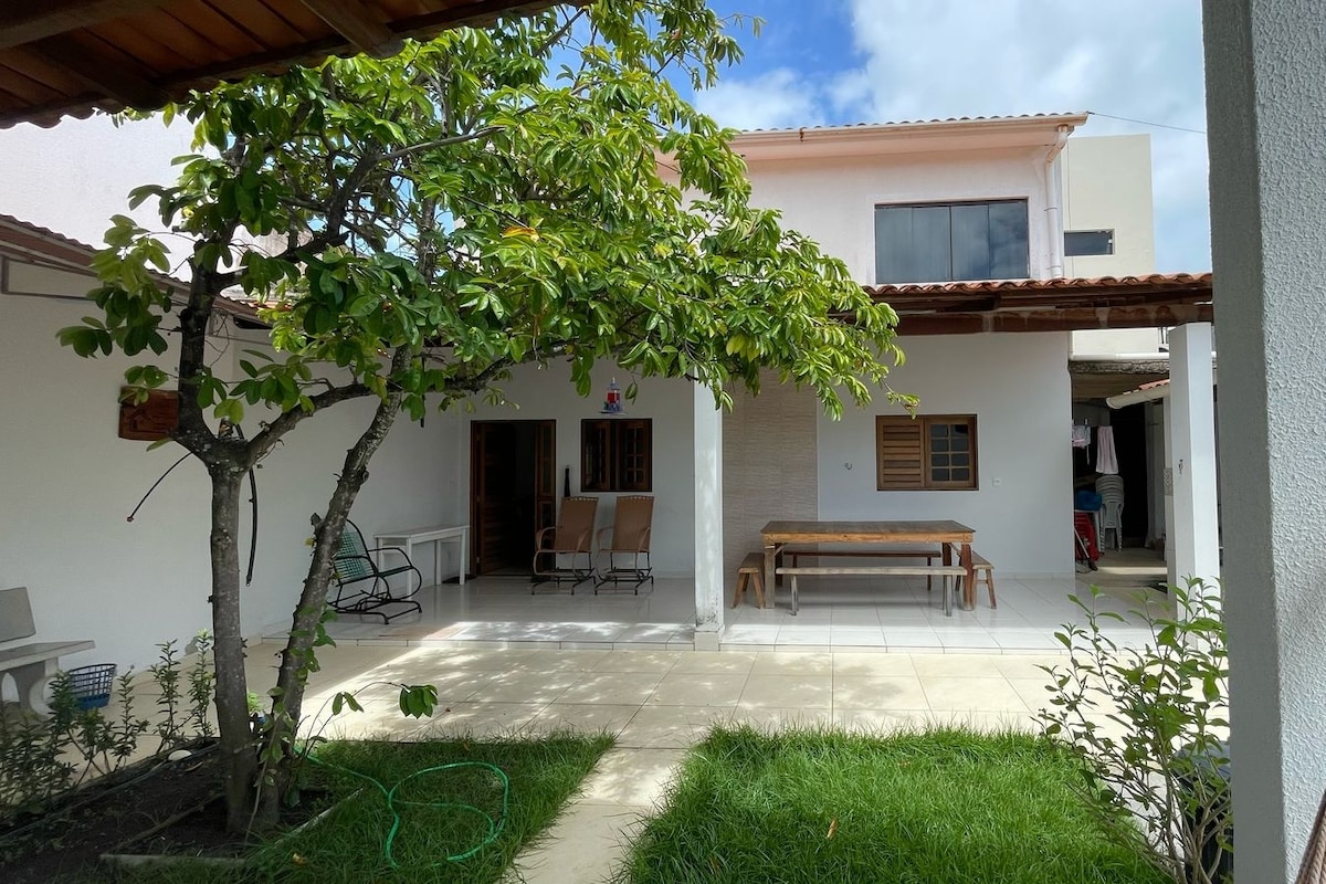 A peaceful outdoor area is depicted, featuring a shaded space with a large wooden dining table and several chairs. Lush green grass surrounds the patio, with a small tree providing additional shade. The home's white walls create a bright and open atmosphere.