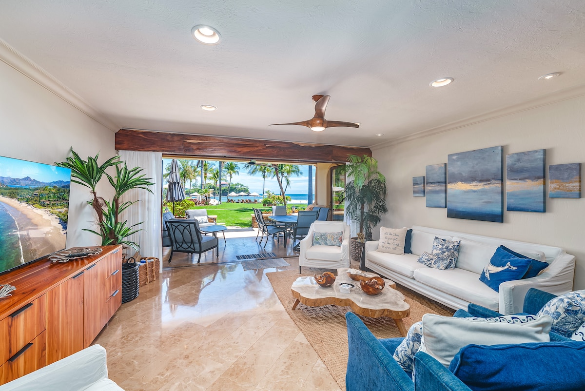A spacious living area features a large flat-screen television and light-colored sofas, complemented by decorative pillows. Large windows frame views of the outdoors, where palm trees can be seen alongside seating on a patio. Natural light illuminates the room, enhancing the relaxed ambiance.